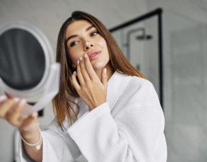 Cuidados com a pele: Mulher de roupão branco, cabelos longos e fios escuros, se olhando em um espelho de mão, observando o próprio rosto enquanto toca na bochecha