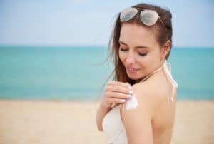 Mulher de pele branca na praia de biquini branco,cabelos escuros soltos, presos apenas pelo óculos escuros na cabeça como uma tiara passando protetor solar no ombro