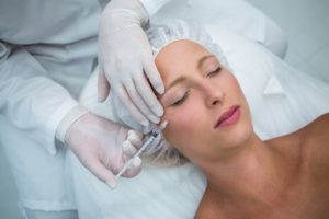 Mulher deitada em uma maca de procedimentos com touca na cabeça enquanto uma dermatologista de luvas descartáveis está segurando uma seringa e aplicando botox próximo ao olho direito da paciente